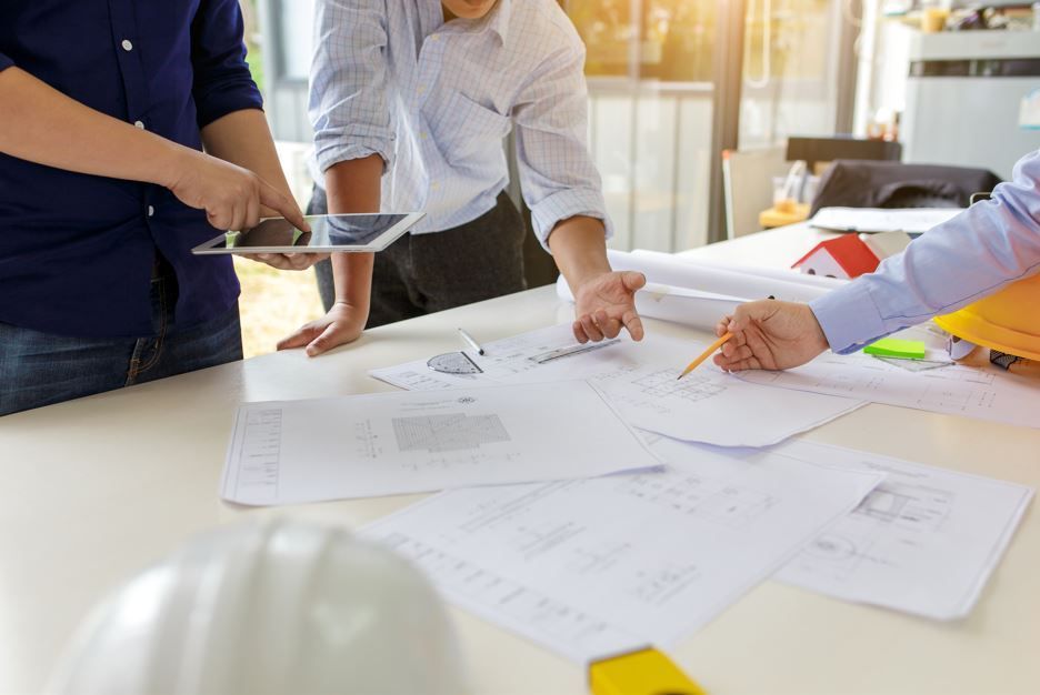 Tres personas en una mesa revisan planos arquitectónicos. Una señala, otra sostiene una tableta, otra lleva un casco.
