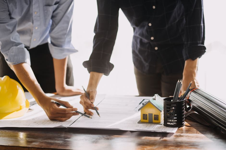 Dos personas revisando planos arquitectónicos en un escritorio, con una casa modelo y herramientas de dibujo.