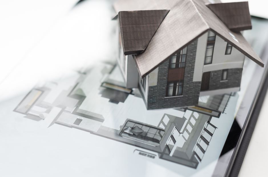 Maqueta de casa en una tableta con planos arquitectónicos. Techo gris, piedra y fachada blanca.