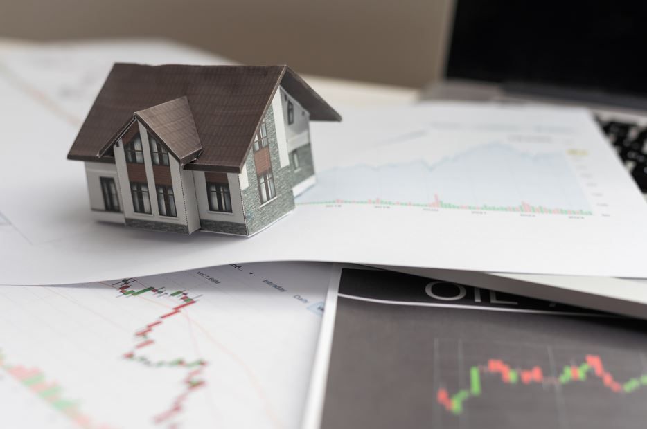 Casa en miniatura sobre documentos con gráficos financieros y una computadora portátil.