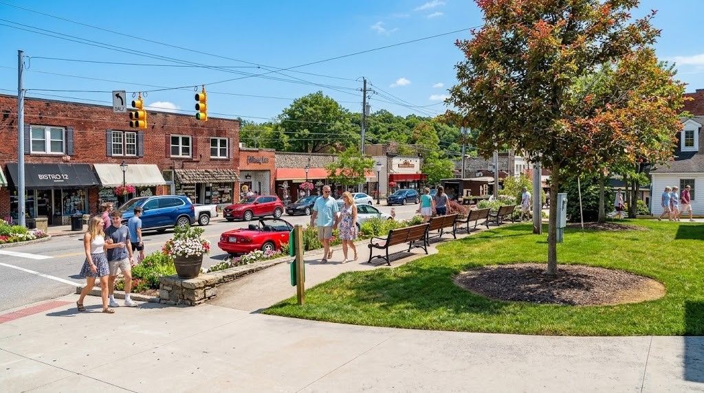 Sunny town square with people walking, shops, parked cars, and a grassy park area with a tree and benches