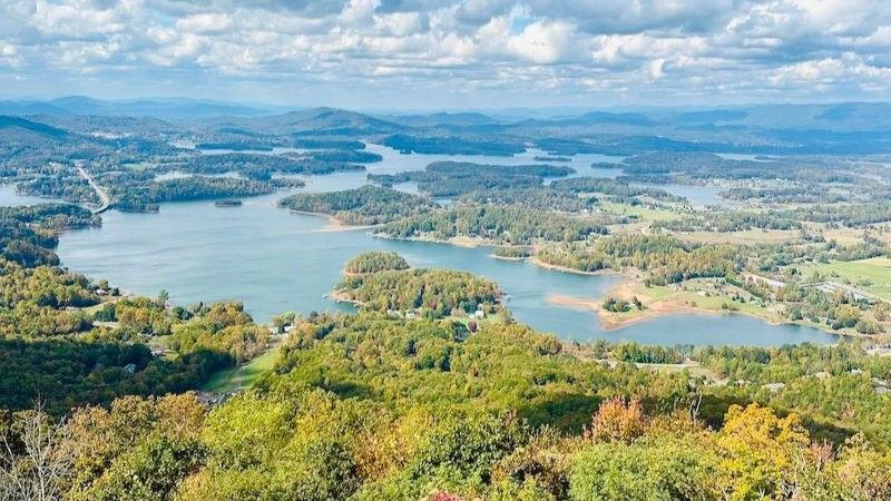 Panoramic lake and rolling hills under a partly cloudy sky, with green forests in the foreground