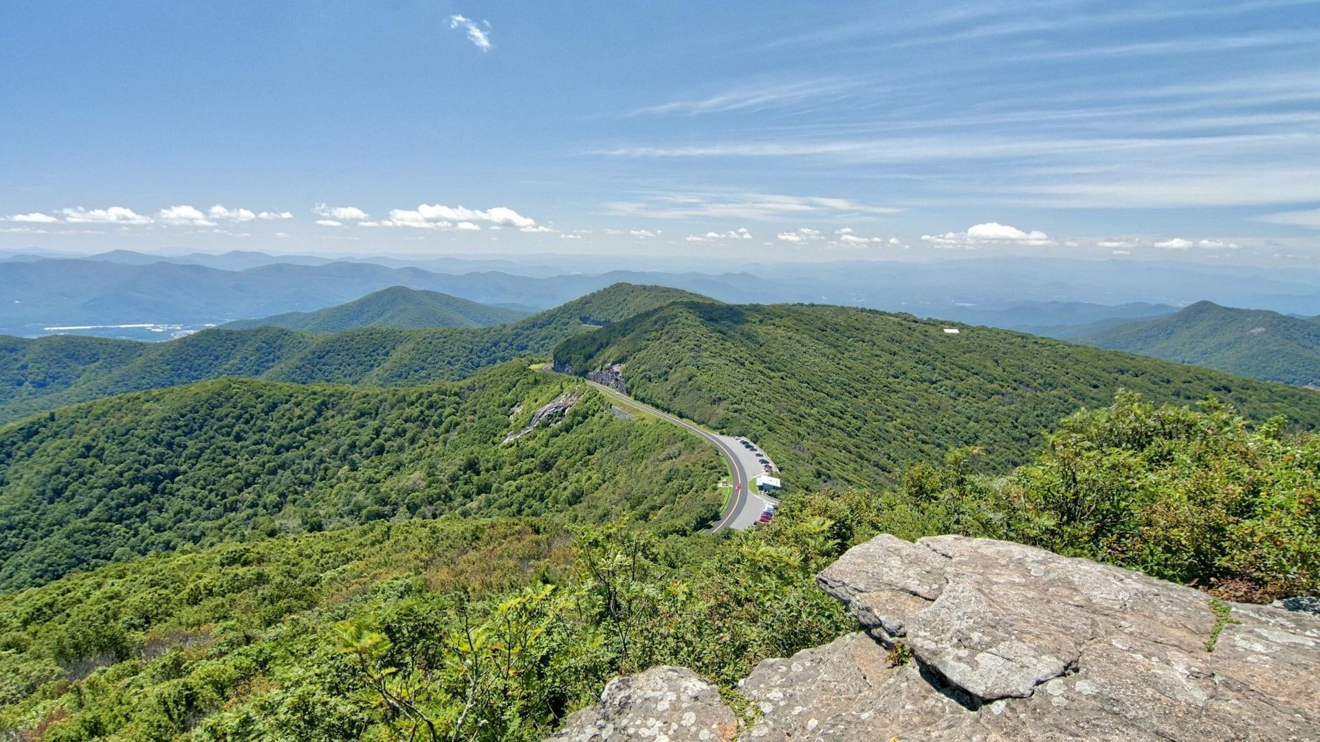 Mountain road winding through green hills under a blue sky, viewed from a rocky overlook