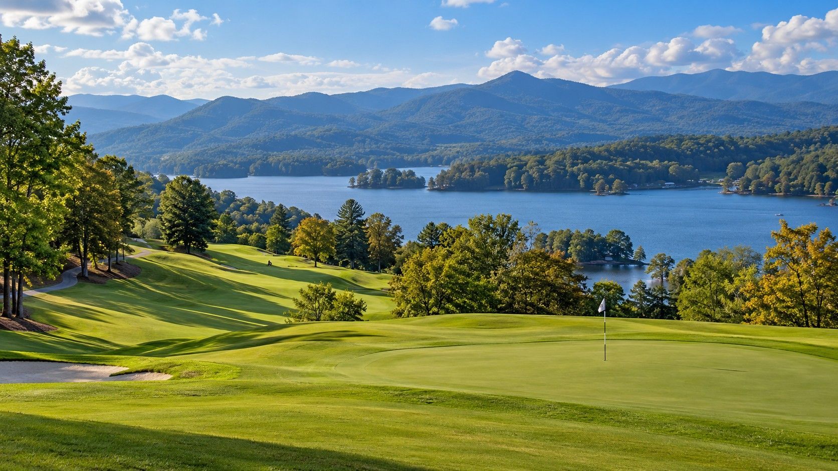 Sunny golf course overlooking a blue lake and forested mountains under a partly cloudy sky