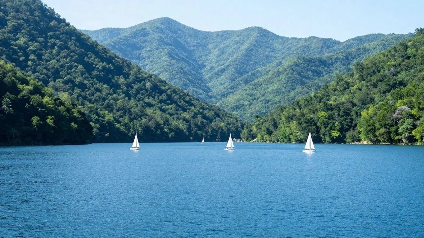Blue lake with three small sailboats, surrounded by green forested mountains under a clear sky