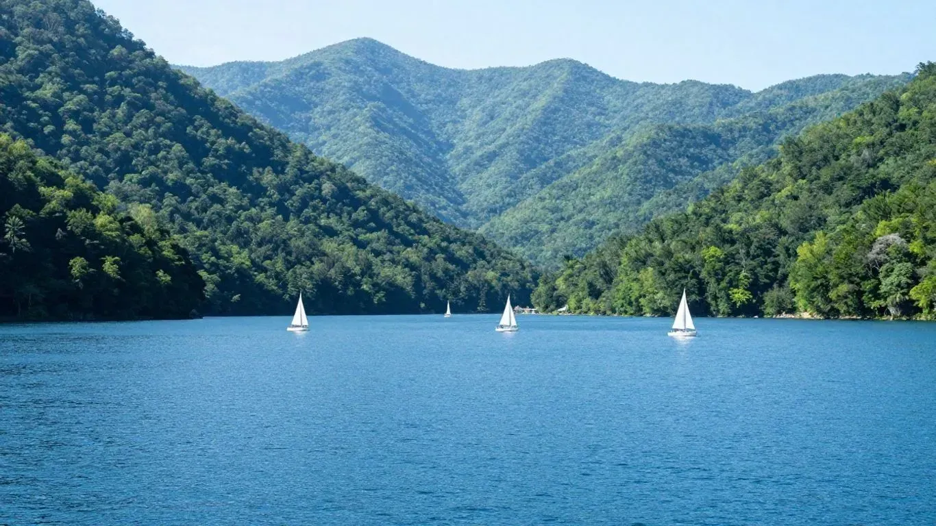 Blue lake with small white sailboats, surrounded by forested mountains under a clear sky