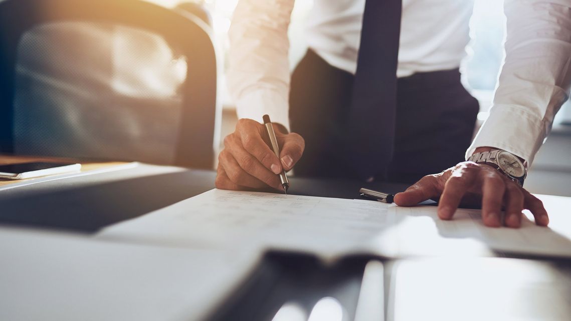 Una persona vestida con camisa blanca y corbata se inclina sobre un escritorio, escribiendo en un documento con un bolígrafo.