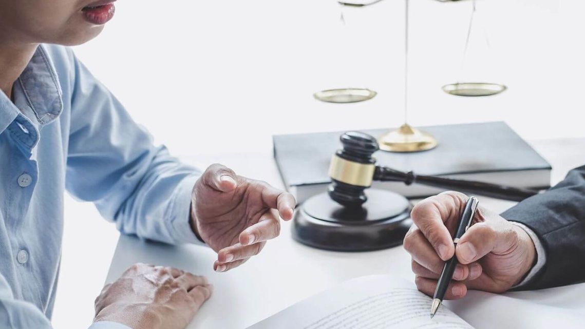 Una persona con camisa azul consulta con un profesional que redacta un documento, con un mazo legal y una balanza al fondo.