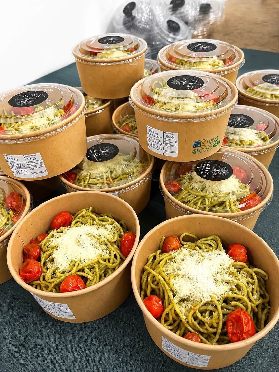 Pila de recipientes para llevar con fideos, adornados con tomate y con tapas transparentes, colocados sobre una mesa