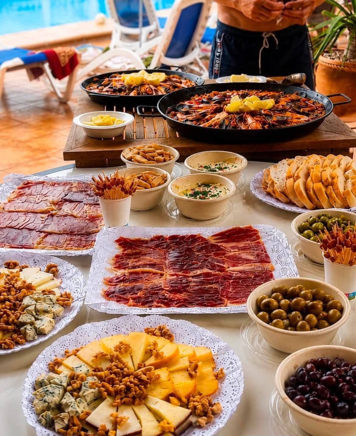 Mesa de buffet con una variedad de platos, embutidos en lonchas, aceitunas y bollería, con un cocinero preparando comida al fondo