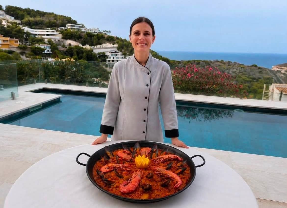 Mujer de pie detrás de una paellera en una terraza con vistas a una piscina y al mar
