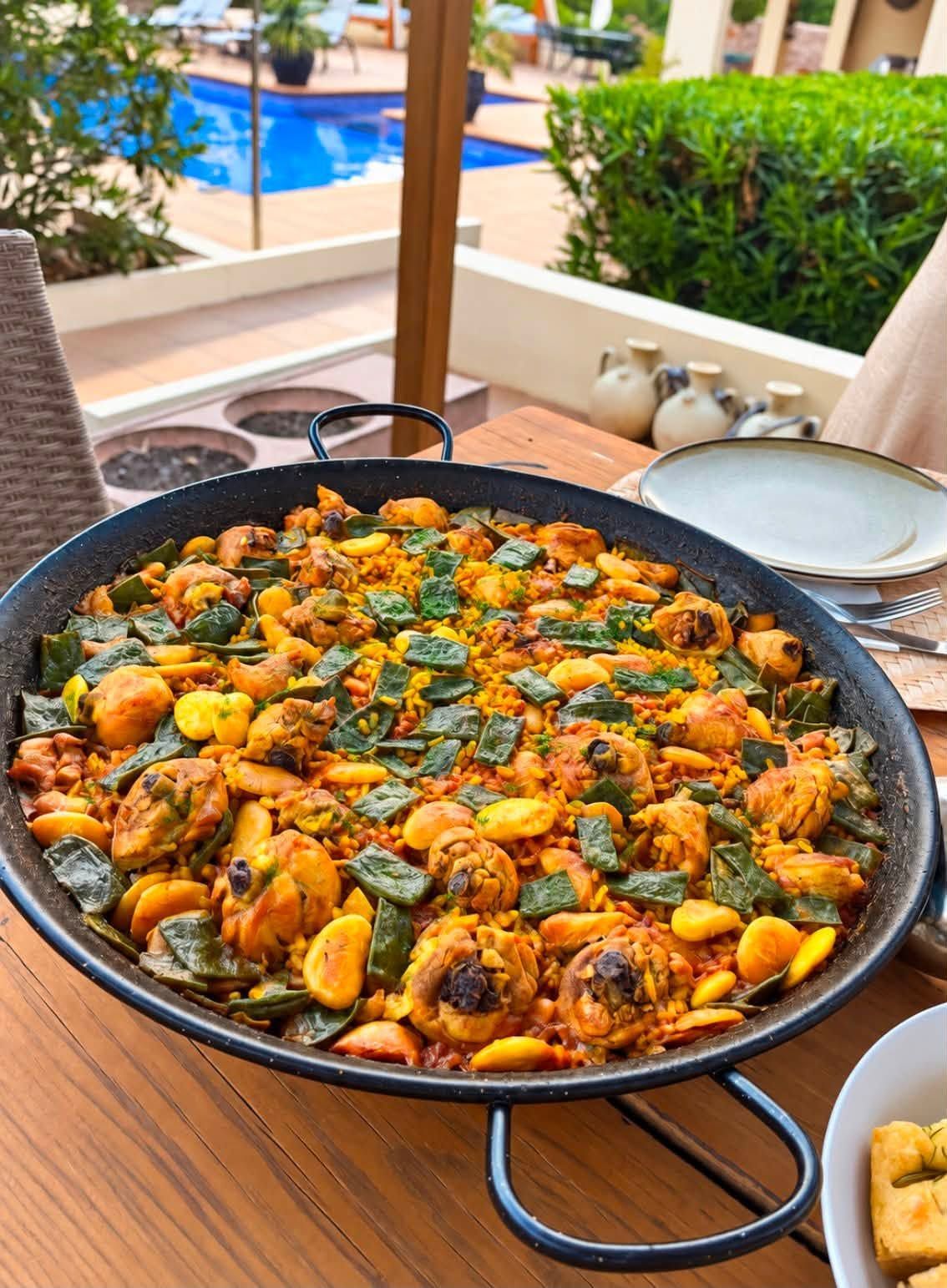 Gran paellera de marisco sobre una mesa de madera junto a una piscina, decorada con hierbas aromáticas y gajos de limón