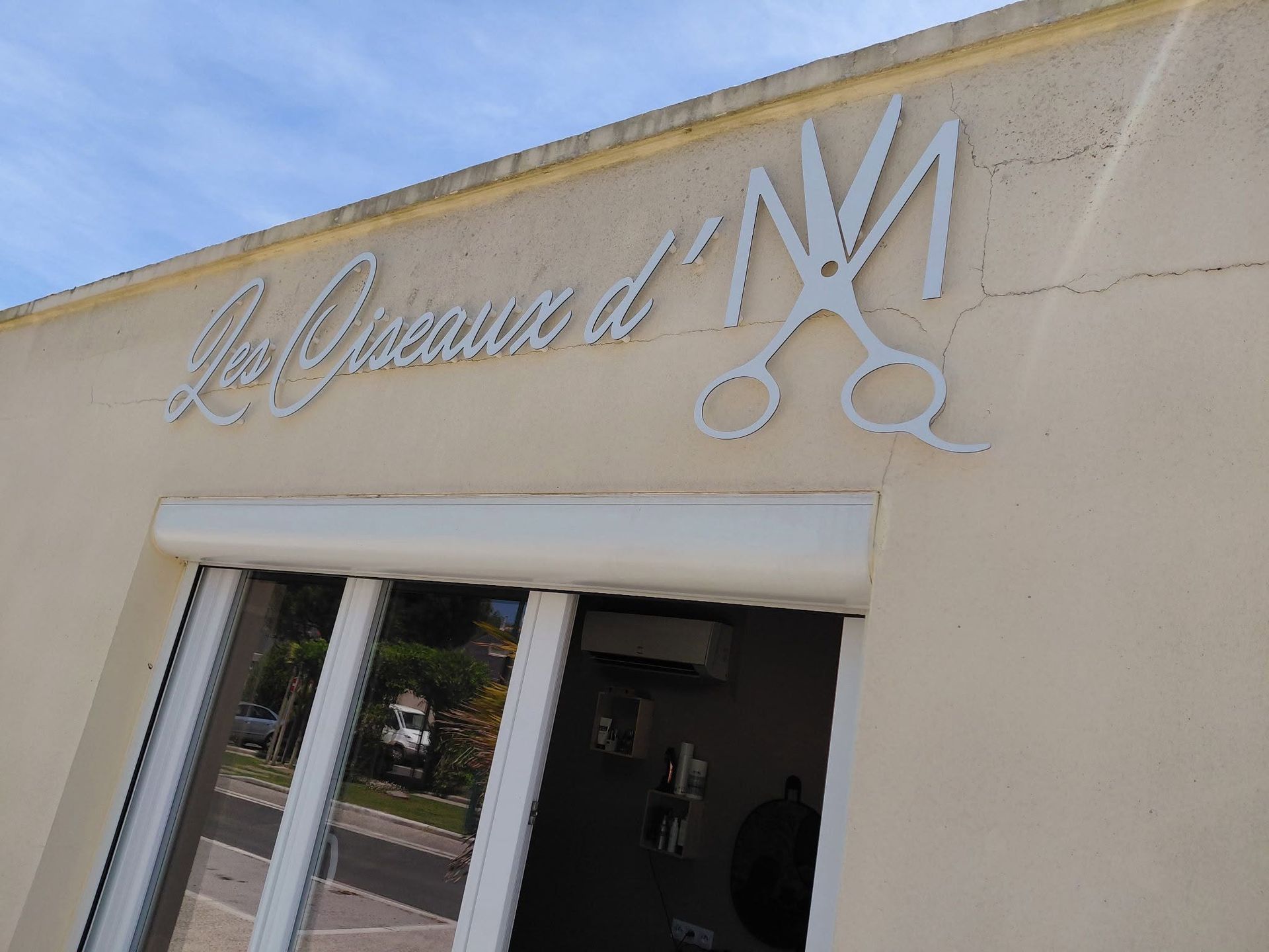 Inscription blanche - Les Ciseaux d'M - en écriture cursive sur la façade beige d'un bâtiment, au-dessus d'une porte vitrée.