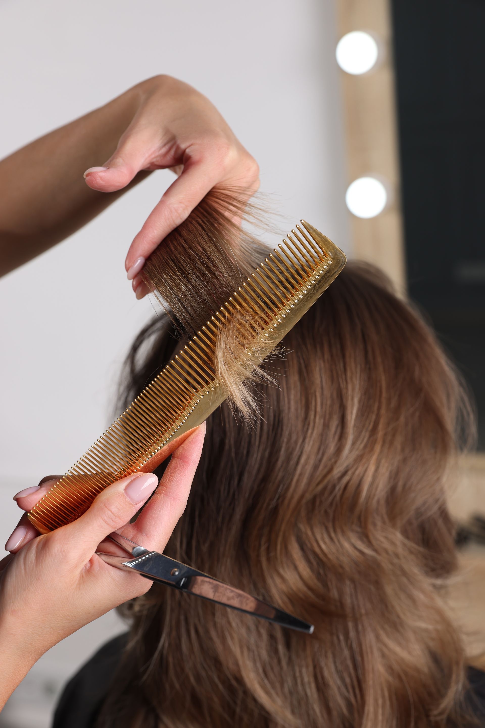 Dans un salon de coiffure, une coiffeuse peigne et coupe des cheveux châtain avec des ciseaux et un peigne doré.