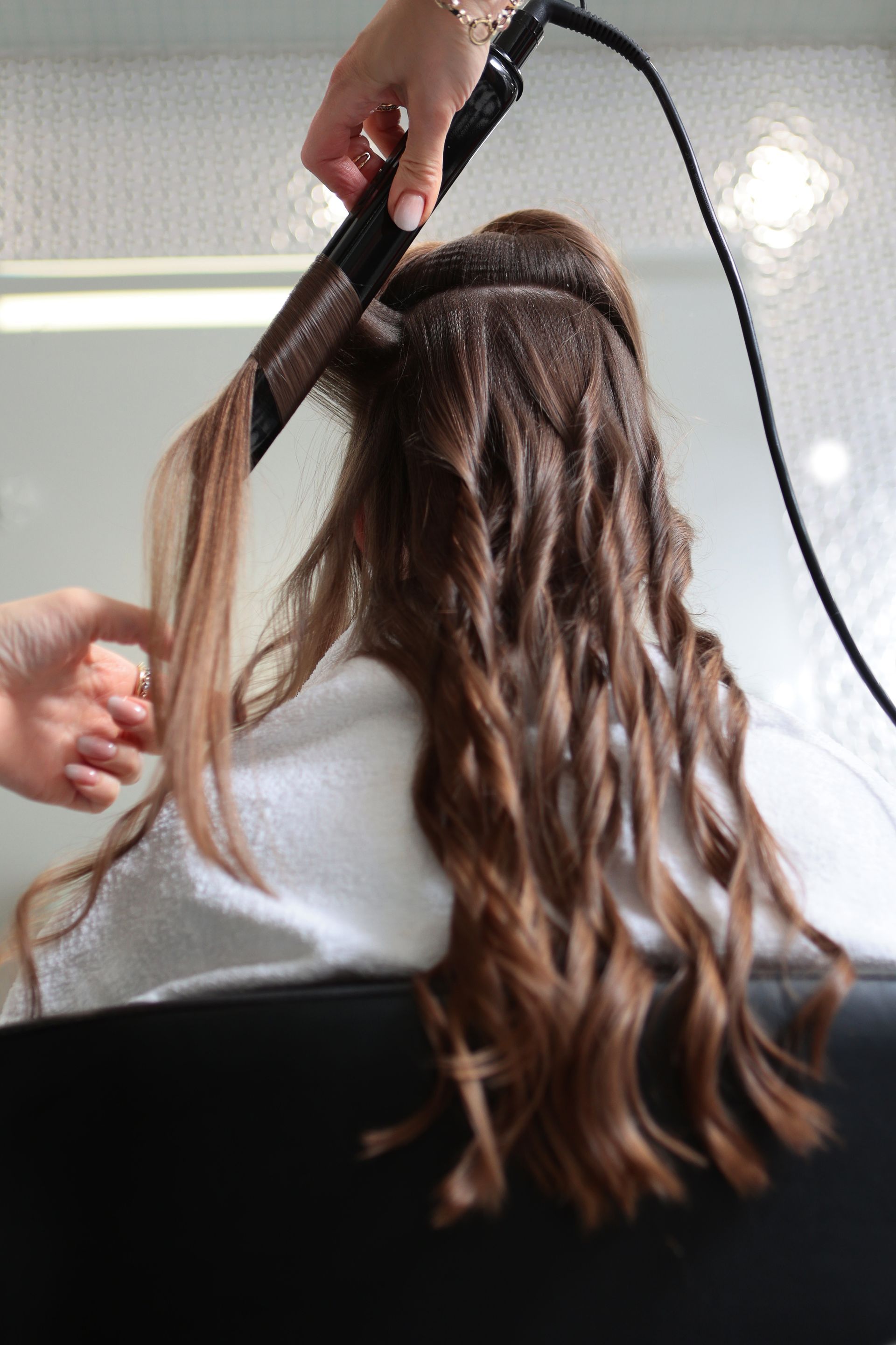 Une coiffeuse boucle de longs cheveux châtains  avec un fer à friser chauffant sur un fauteuil de salon