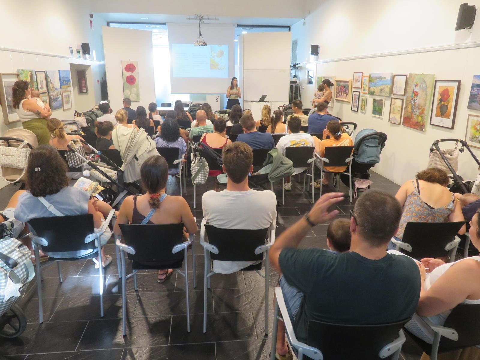 El público se sienta en una sala luminosa frente a una pantalla de presentación y un orador situado al frente.