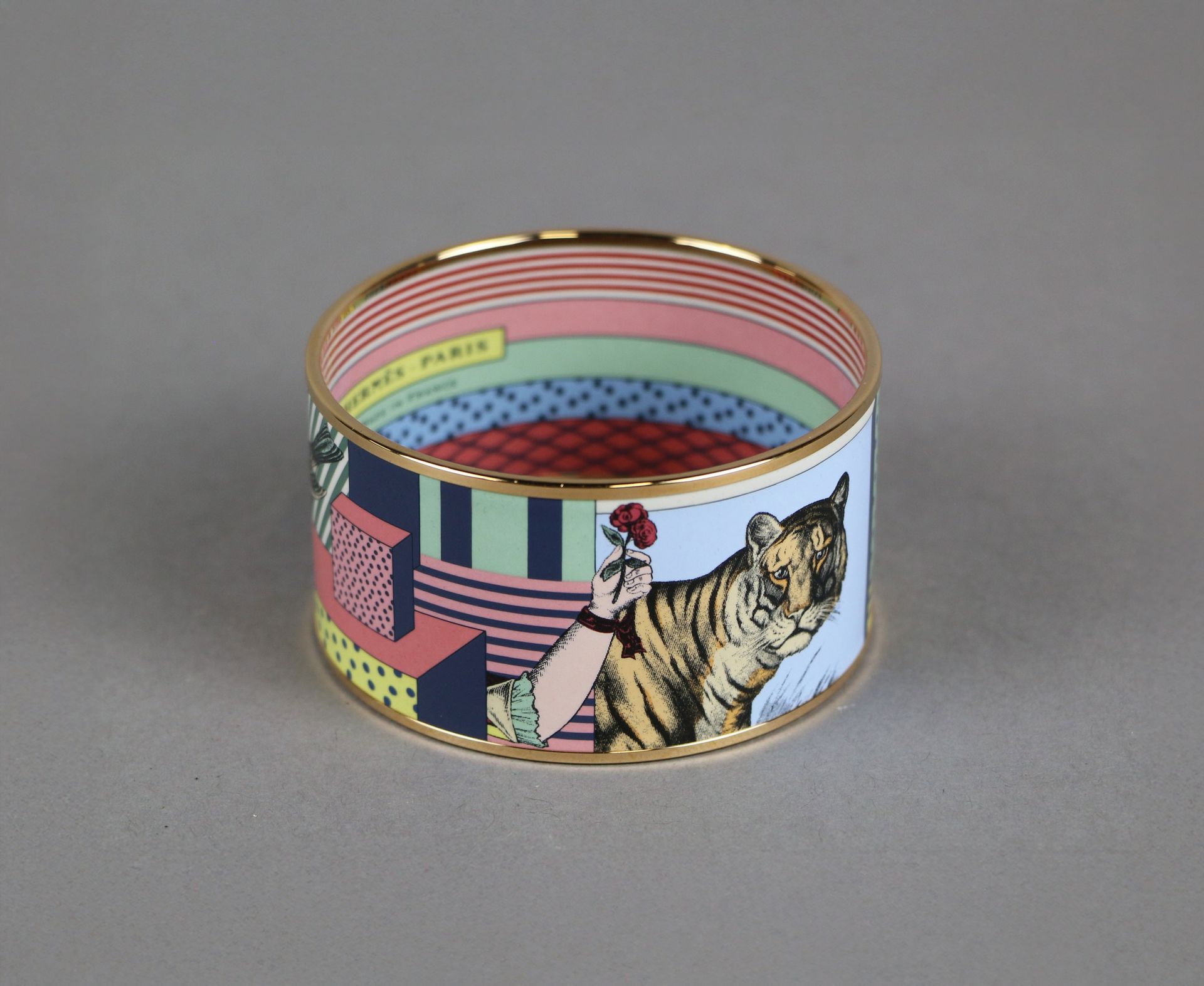 Un bracelet Hermès à bord doré avec une illustration de tigre et un motif floral sur fond gris.