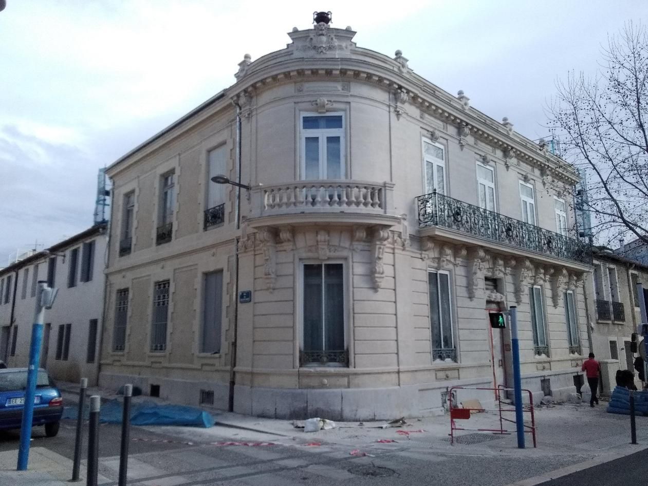 Un immeuble d'angle néoclassique couleur crème, avec une façade arrondie, un balcon orné et des fenêtres cintrées, sous un ciel gris.