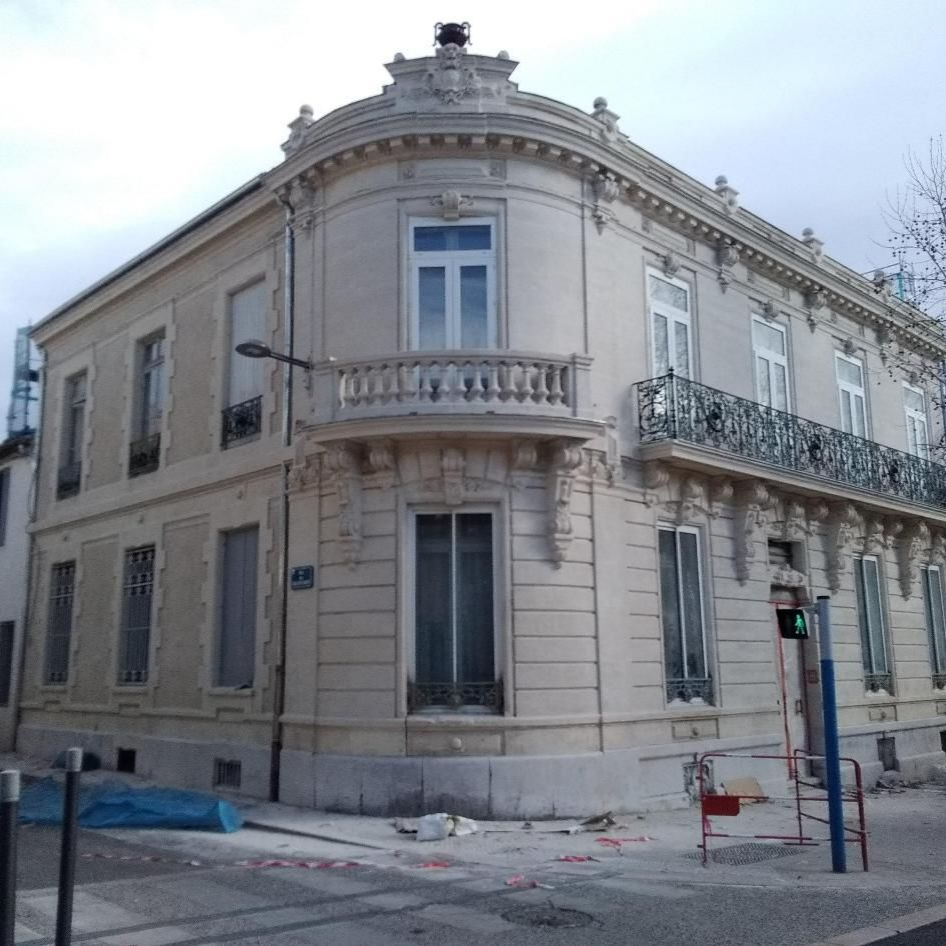 Un immeuble de deux étages, de couleur crème, situé à un angle de rue, avec des balcons ornés et une façade arrondie.