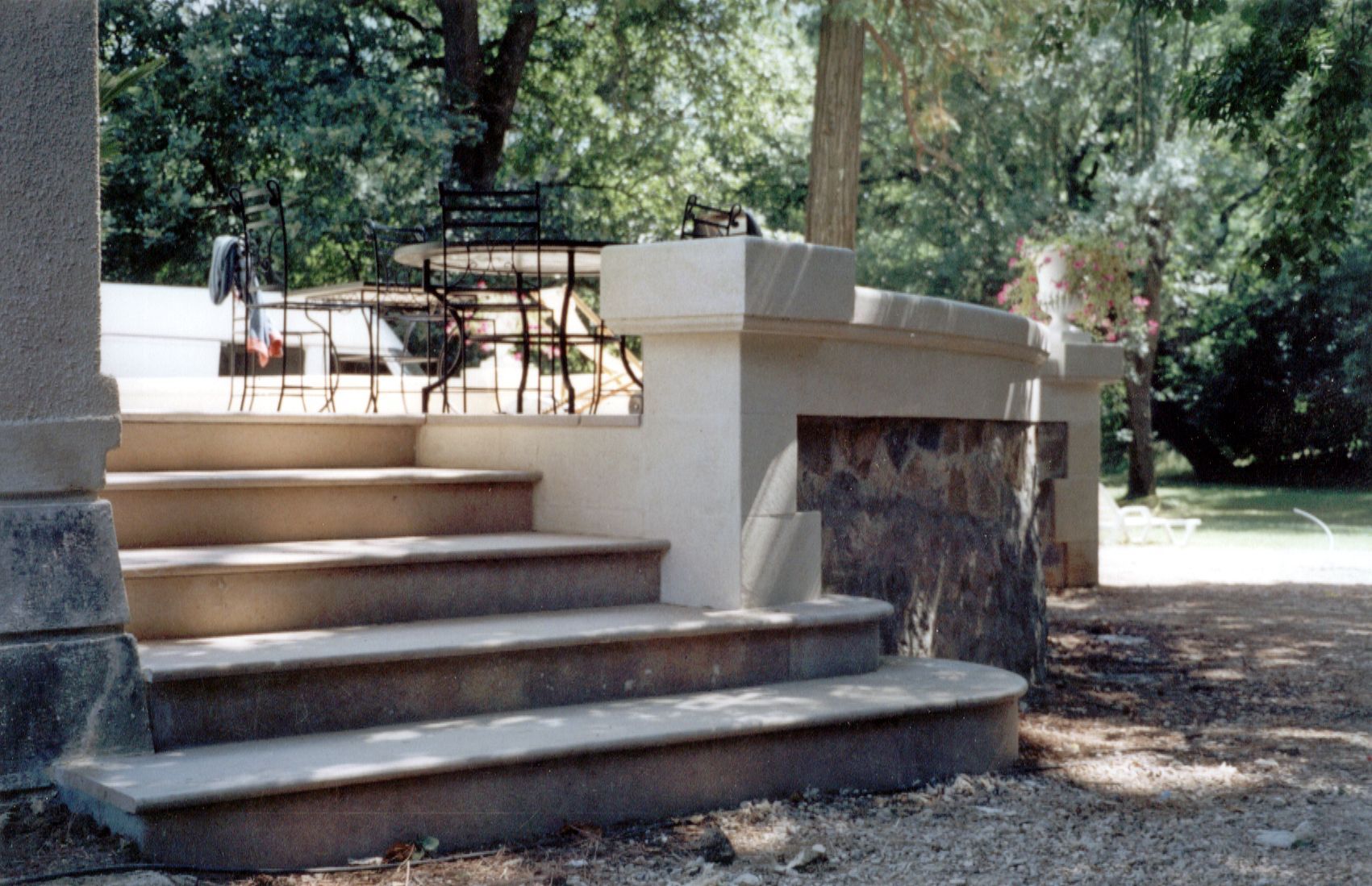 Des marches en béton agrémentées de pierres mènent à une terrasse aménagée avec du mobilier de jardin, dans un cadre champêtre.