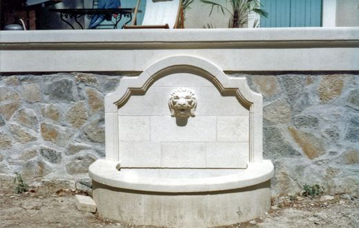 Une fontaine murale en pierre ornée d'un bec verseur en forme de tête de lion sculptée, adossée à un mur de soutènement en pierre dans un espace extérieur.