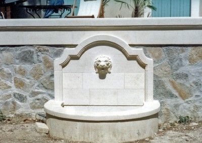 Fontaine en pierre avec un bec en forme de tête de lion encastré dans un mur blanc et gris extérieur