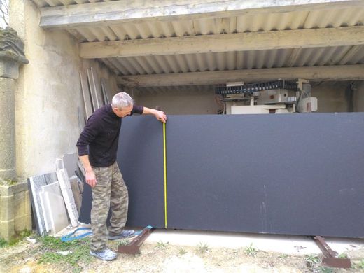 Un homme mesure un grand panneau sombre avec un mètre ruban dans un atelier extérieur.