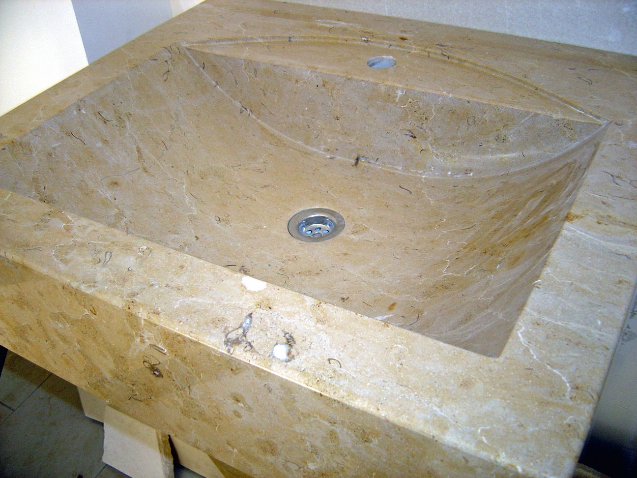 Un lavabo de salle de bains en marbre beige avec une vasque carrée inclinée et un trou d'évacuation central.