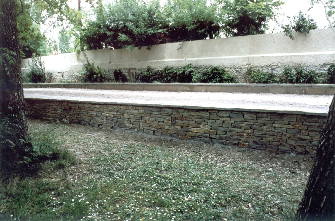Un long terrain de pétanque ou de bocce surélevé, en pierre avec une surface en gravier, situé dans une cour ombragée par des arbres.