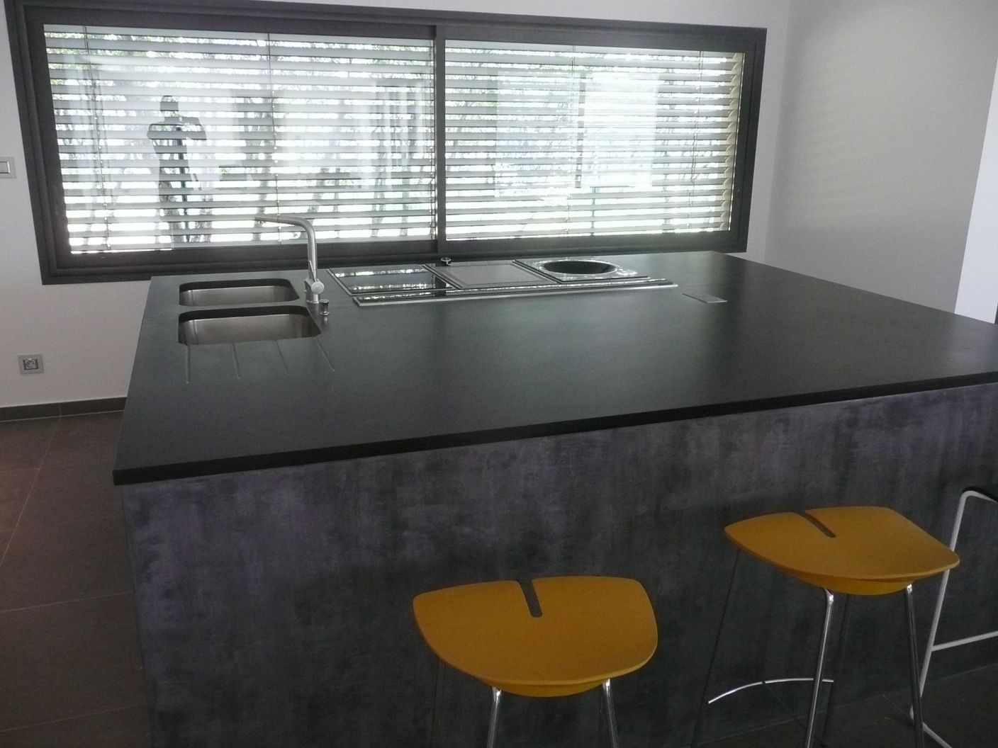 Un îlot de cuisine moderne avec un plan de travail foncé mat, un évier et une plaque de cuisson intégrés, et deux tabourets jaunes dans une pièce lumineuse.