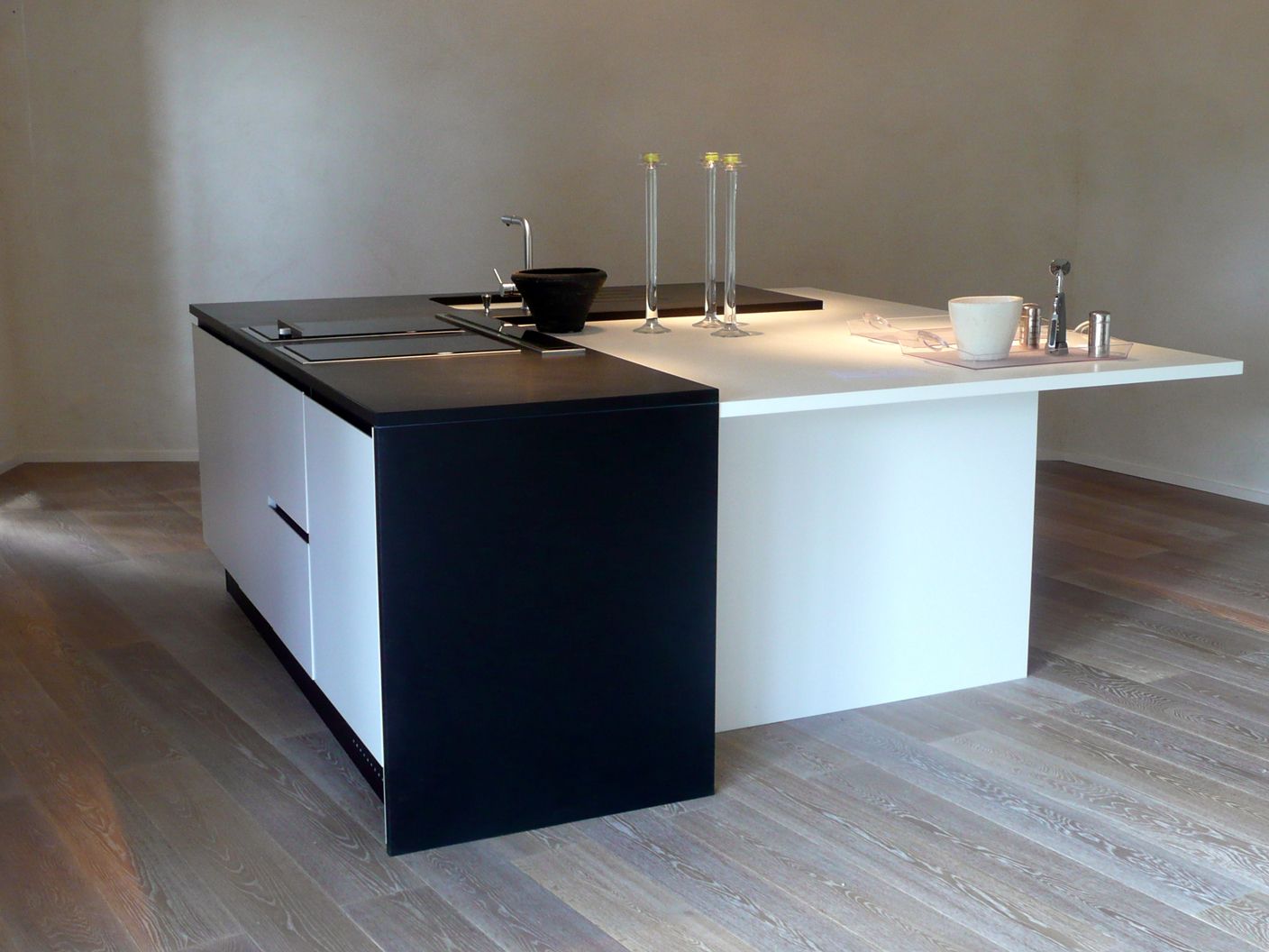 Un îlot de cuisine à deux niveaux, avec un plan de travail noir et un socle blanc, installé sur un parquet clair dans une pièce minimaliste.