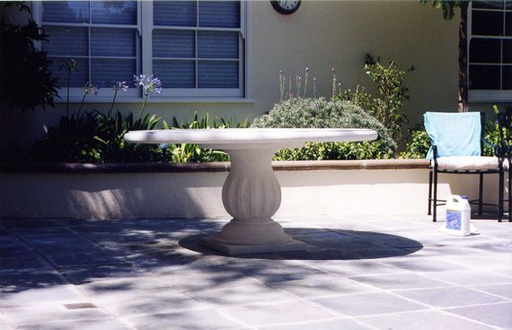 Table de jardin en pierre sur une terrasse ensoleillée, à côté d'une chaise et d'une bouteille de produit nettoyant.