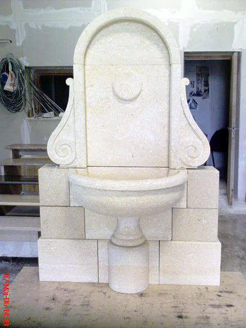Évier en pierre blanche sculptée avec dosseret arqué, adossé à un mur d'atelier en blocs de pierre empilés.