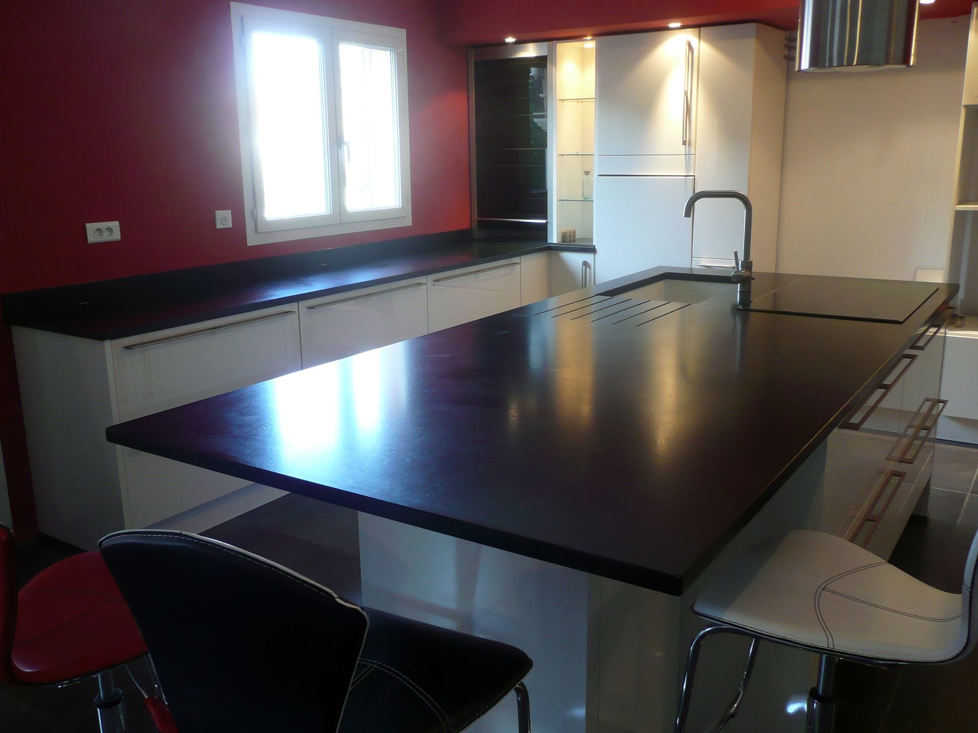 Cuisine moderne avec un grand îlot noir, des placards blancs, des murs rouges et une fenêtre.