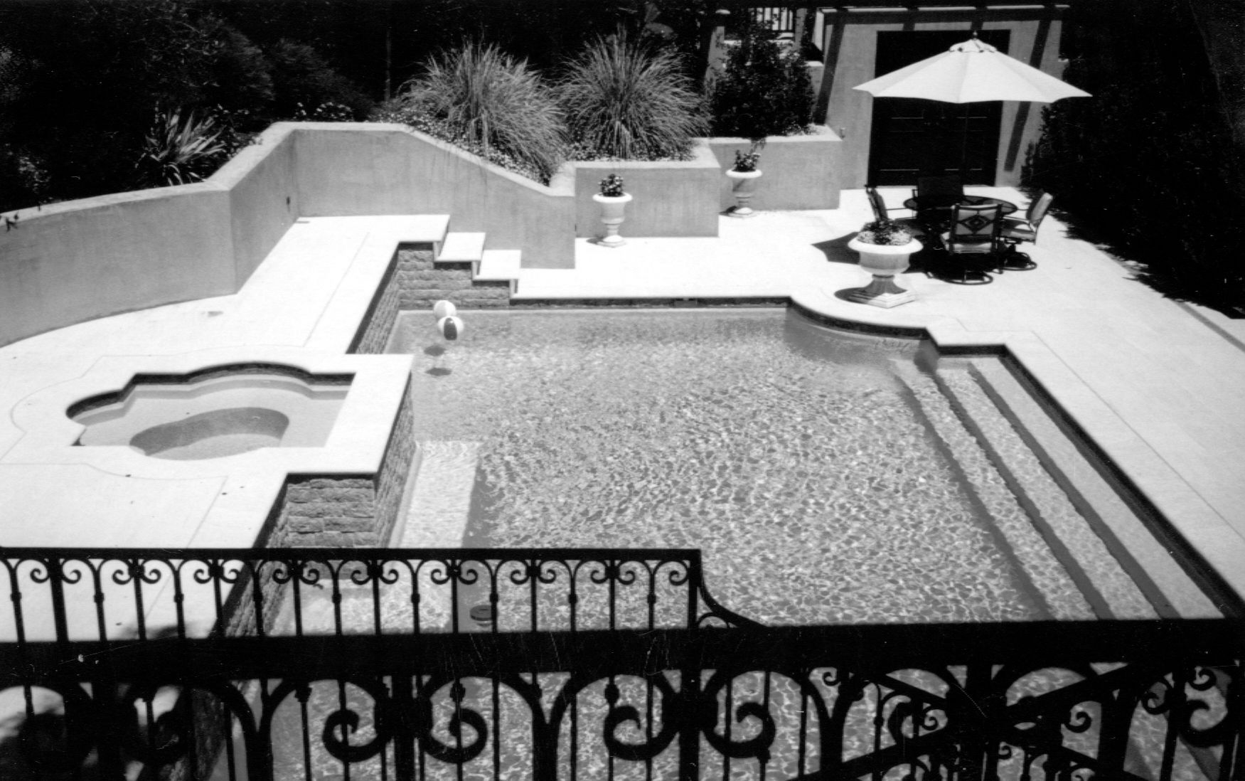 Vue en plongée, en noir et blanc, d'une piscine avec jacuzzi attenant et d'une terrasse aménagée avec des sièges extérieurs.