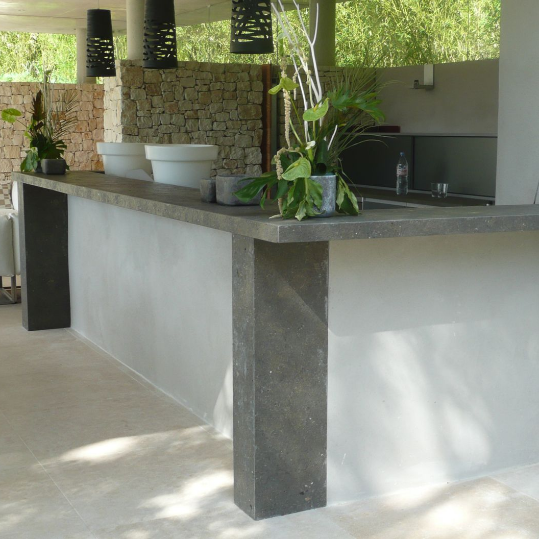 Un comptoir de bar en pierre à la surface grise lisse, des supports d'angle assortis et des plantes en pot dans un cadre extérieur.