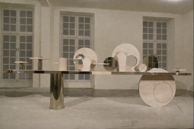 Installation de sculpture abstraite dans une galerie, avec des formes métalliques circulaires sur une longue table réfléchissante.