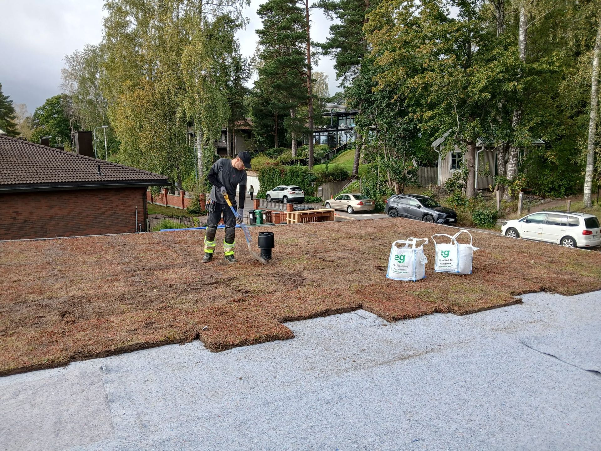Työntekijä levittää katteeksi käytettävää haketta pihalle ajotien viereen. Taustalla näkyy materiaalipusseja ja puita