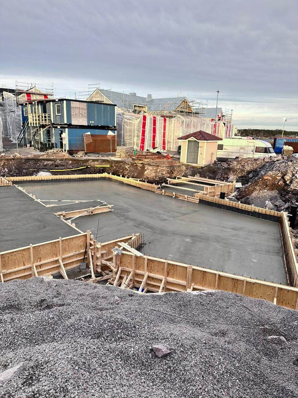 Rakennustyömaa, jolla on betoniperustus, puiset muotit ja materiaaleja levällään teollisuusrakennusten vieressä.