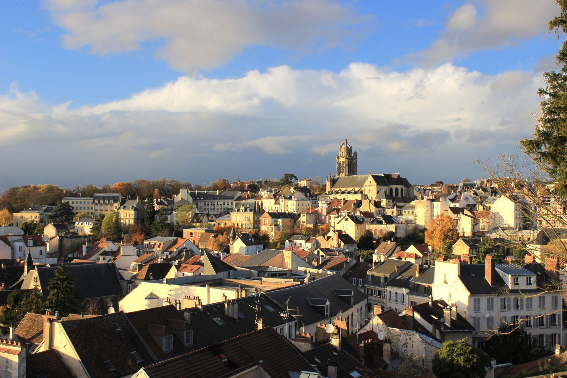 Paysage urbain avec clocher sous un ciel bleu nuageux, et feuillage d'automne au premier plan.