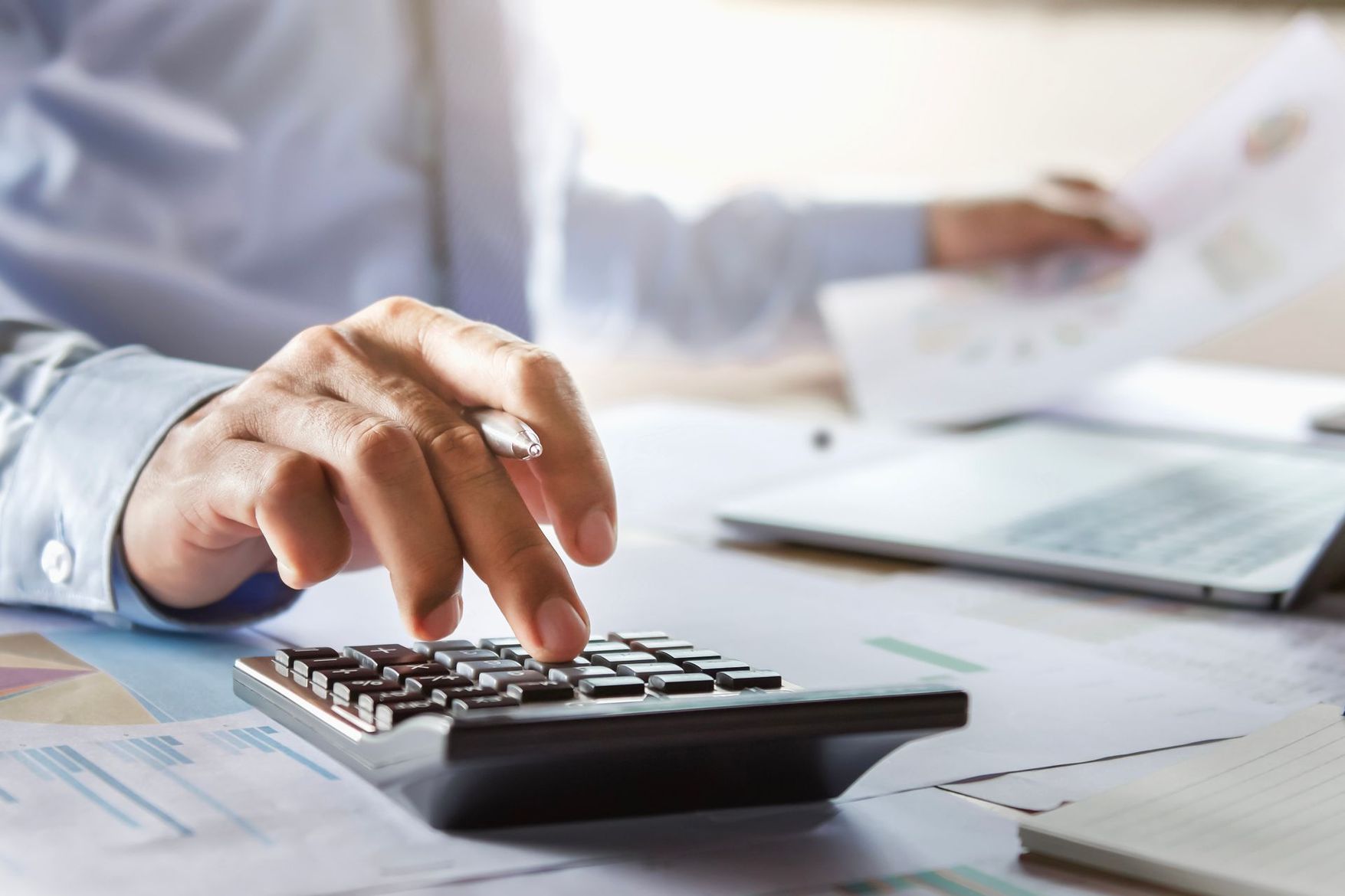 La main d'une personne utilisant une calculatrice, avec des papiers et un ordinateur portable en arrière-plan, concentrée sur des calculs financiers.