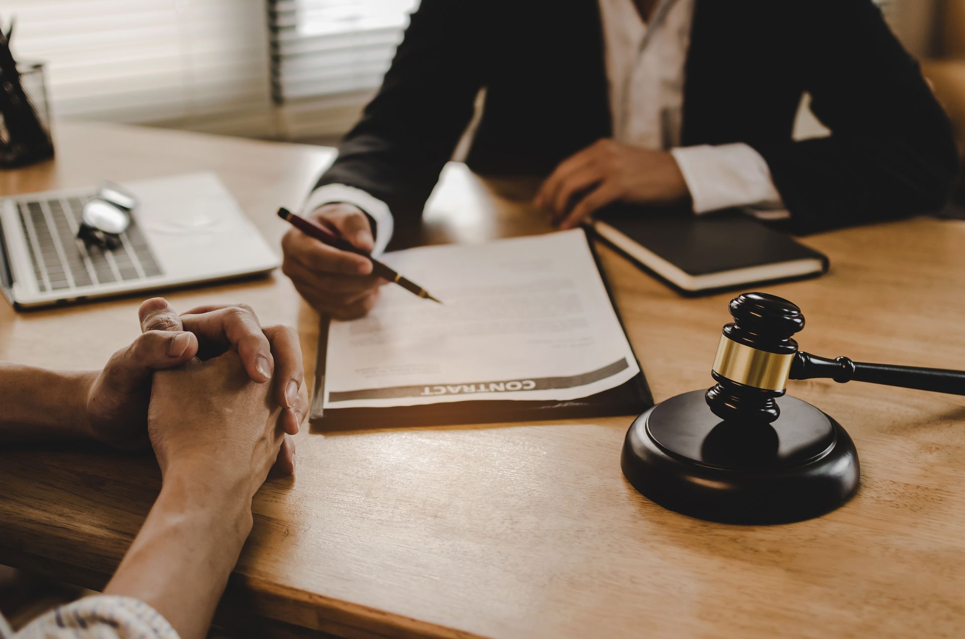 L'avocat désigne un document. Le client a les mains jointes. Un maillet est posé sur la table. Un carnet, un ordinateur portable. Ambiance de bureau.