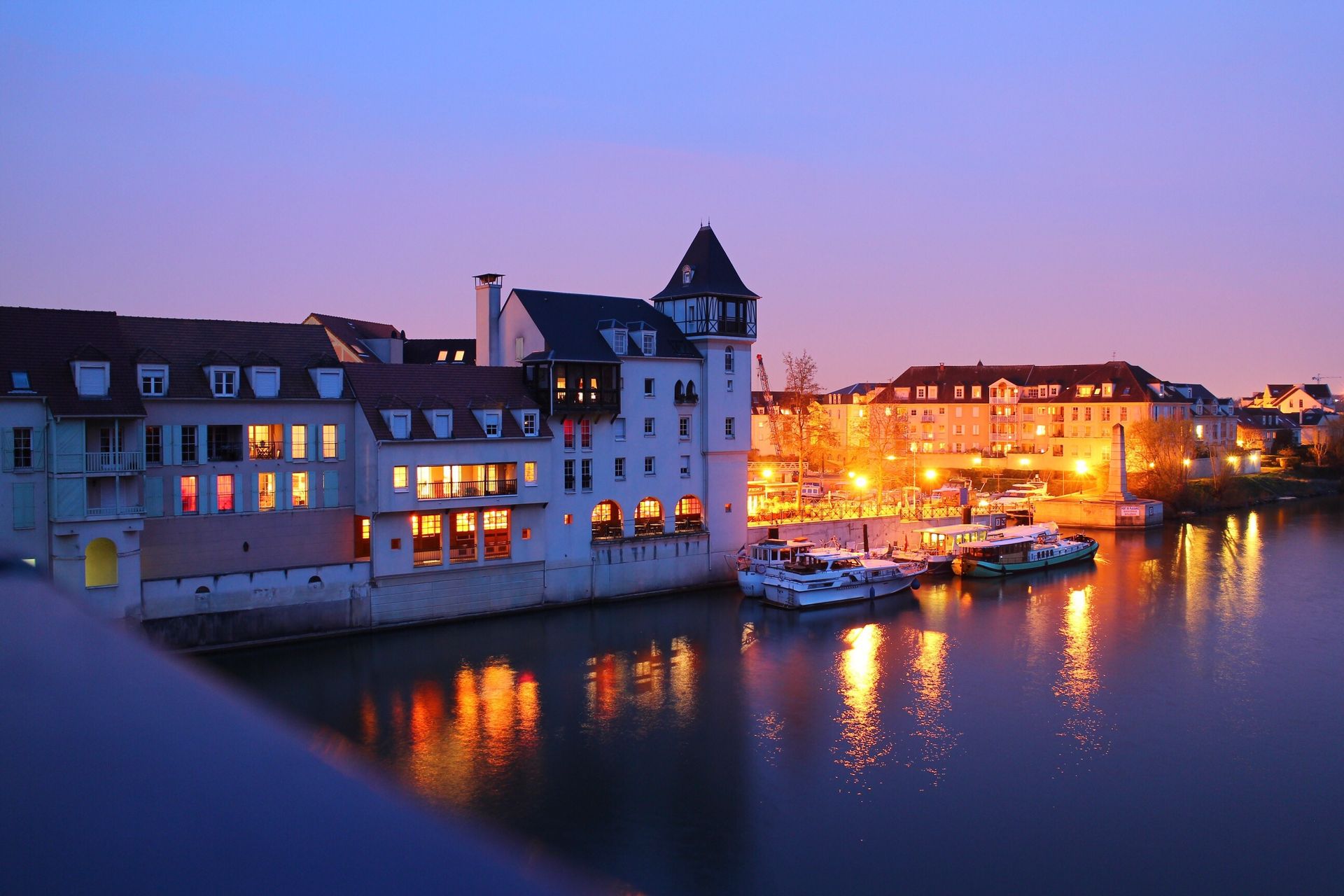 Au crépuscule, des bâtiments bordent une rivière, leurs lumières se reflétant dans l'eau. Des bateaux sont à quai.