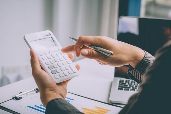 Une personne utilisant une calculatrice, tenant un stylo et travaillant sur des documents près d'un ordinateur portable.