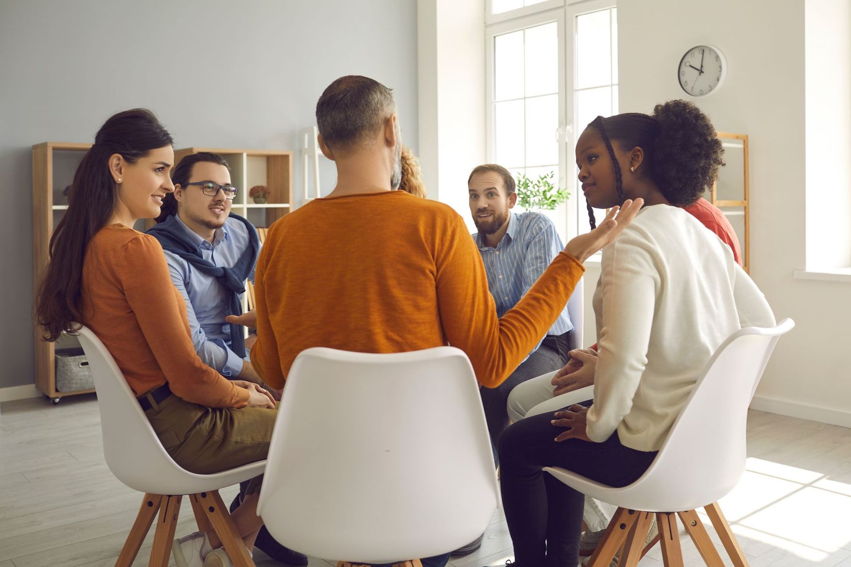 Des personnes assises en cercle, engagées dans une conversation, à l'intérieur d'une pièce avec une fenêtre, discutant ou partageant.