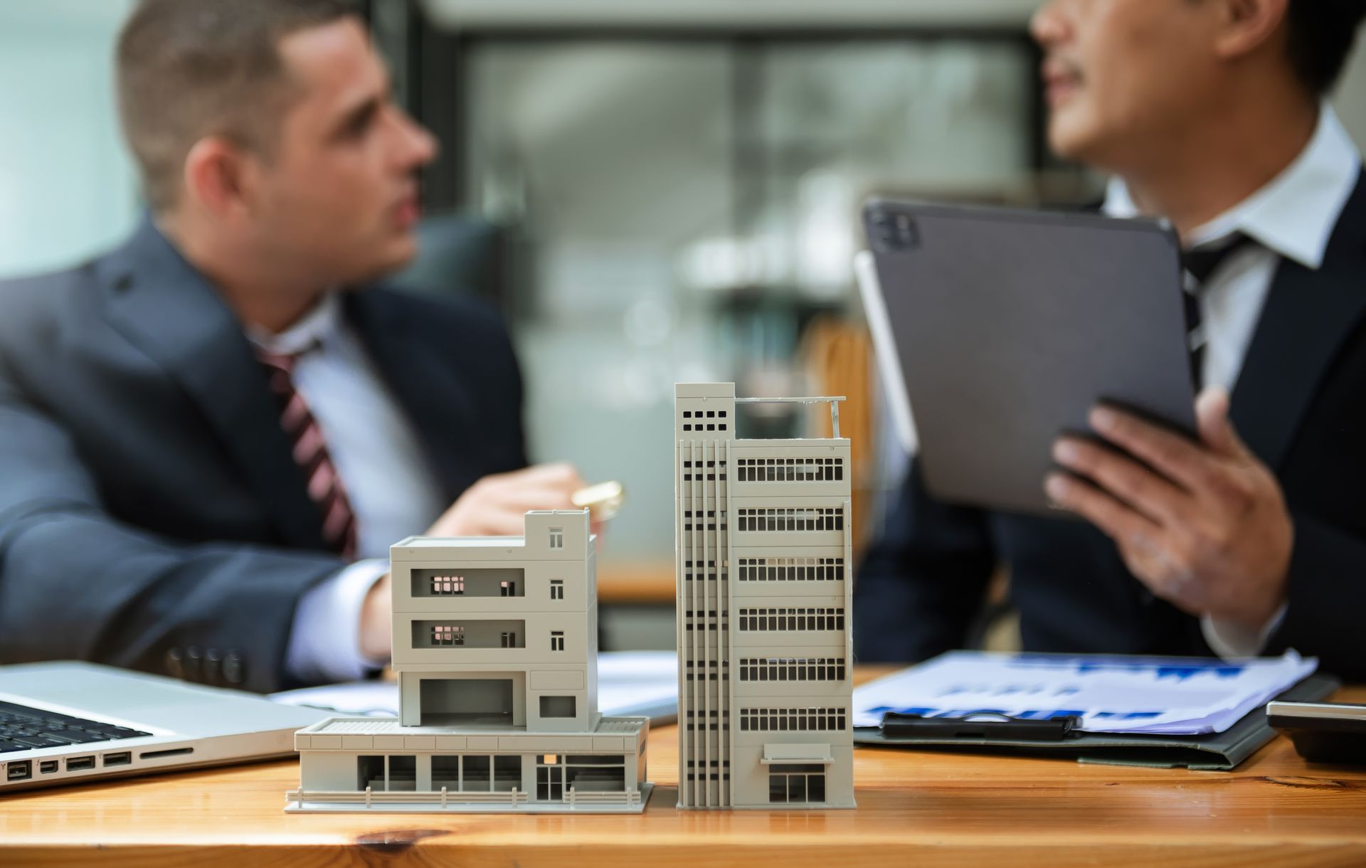 Deux hommes en costume, des maquettes de bâtiments sur un bureau, discutent d'un projet.