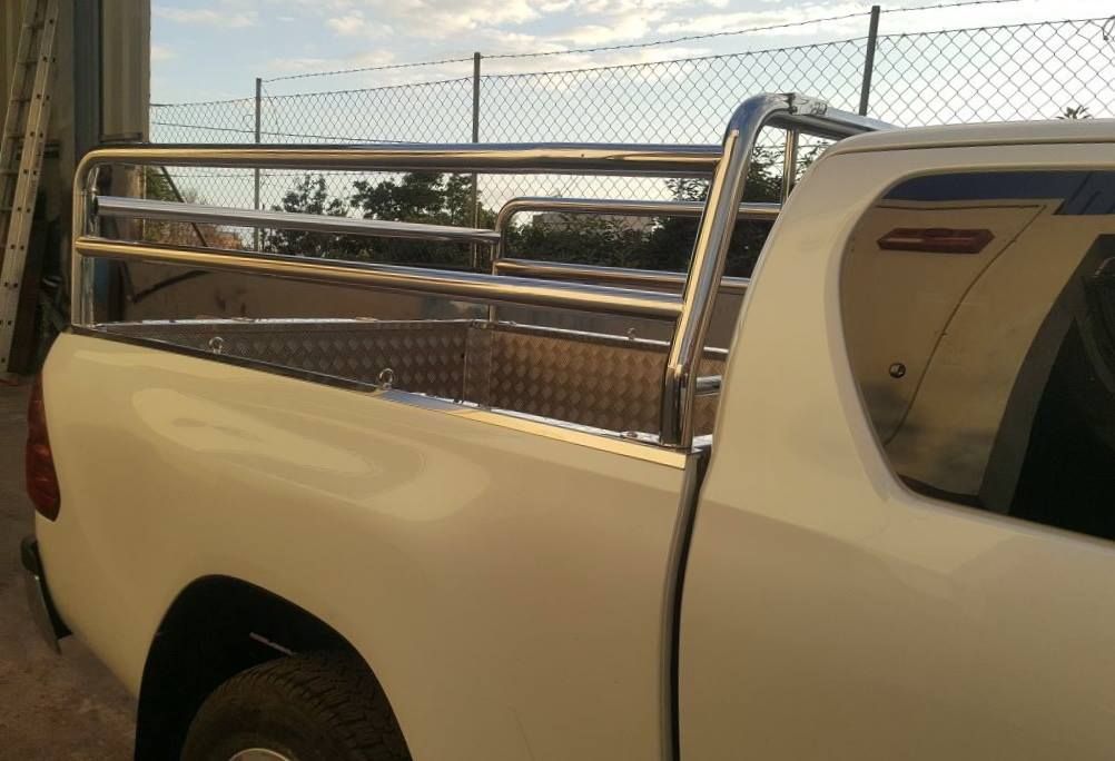 Un camión blanco con una barandilla de acero inoxidable en la parte trasera está estacionado frente a una cerca de alambre.