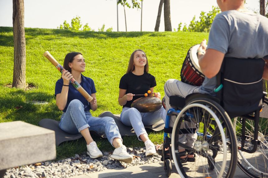 Tres personas tocando instrumentos en un parque: una en silla de ruedas sobre un tambor, dos en el suelo con percusión.