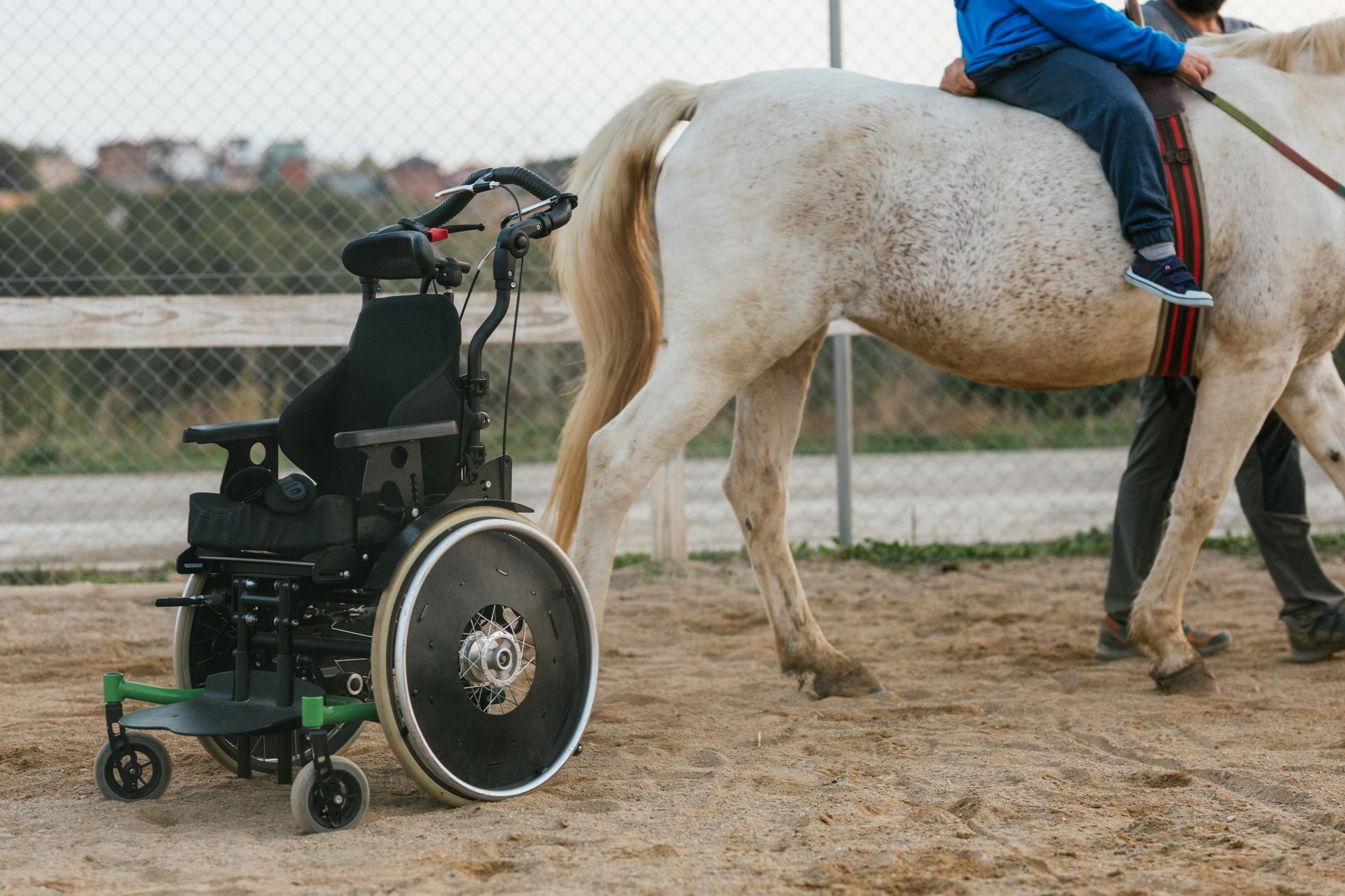 Silla de ruedas junto a un caballo blanco con un niño jinete en un picadero al aire libre.
