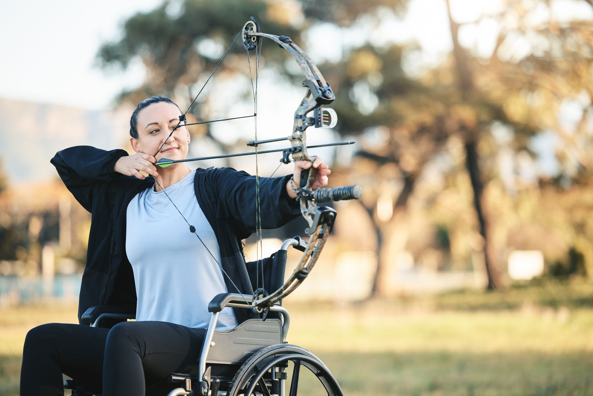 Mujer en silla de ruedas apunta con arco y flecha al aire libre.